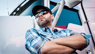 Confident Caucasian male american trucker next to his big rig. Concept of trucking owner operators with man in plaid shirt and baseball cap. Transportation industry professional 18 wheeler driver.